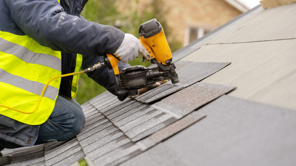 roofer in Tulsa worker in special protective work wear and gloves, using air or pneumatic nail gun and installing asphalt or bitumen shingle on top of the new roof under construction residential building
